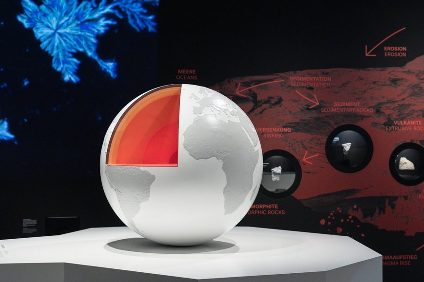 Im Vordergrund steht ein angeschnittenes Modell der Erdkugel, im Hintergrund werden auf einer Infotafel unterschiedliche Prozesse von Vulkangesteinen skizziert