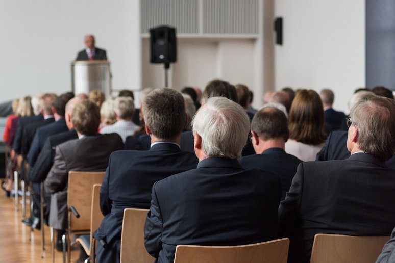Frontalaufnahme eines vollen Saals, gefüllte Sitzreihen sind dem Sprecher am Podium zugewandt.