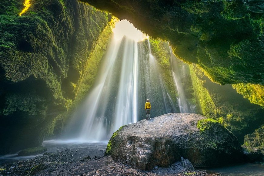 Durch eine Öffnung in der Decke strömen Lichtstrahlen und Wasser in die moosbewachsene Höhle. Eine Person steht auf einem Felsen und blickt der Öffnung entgegen.